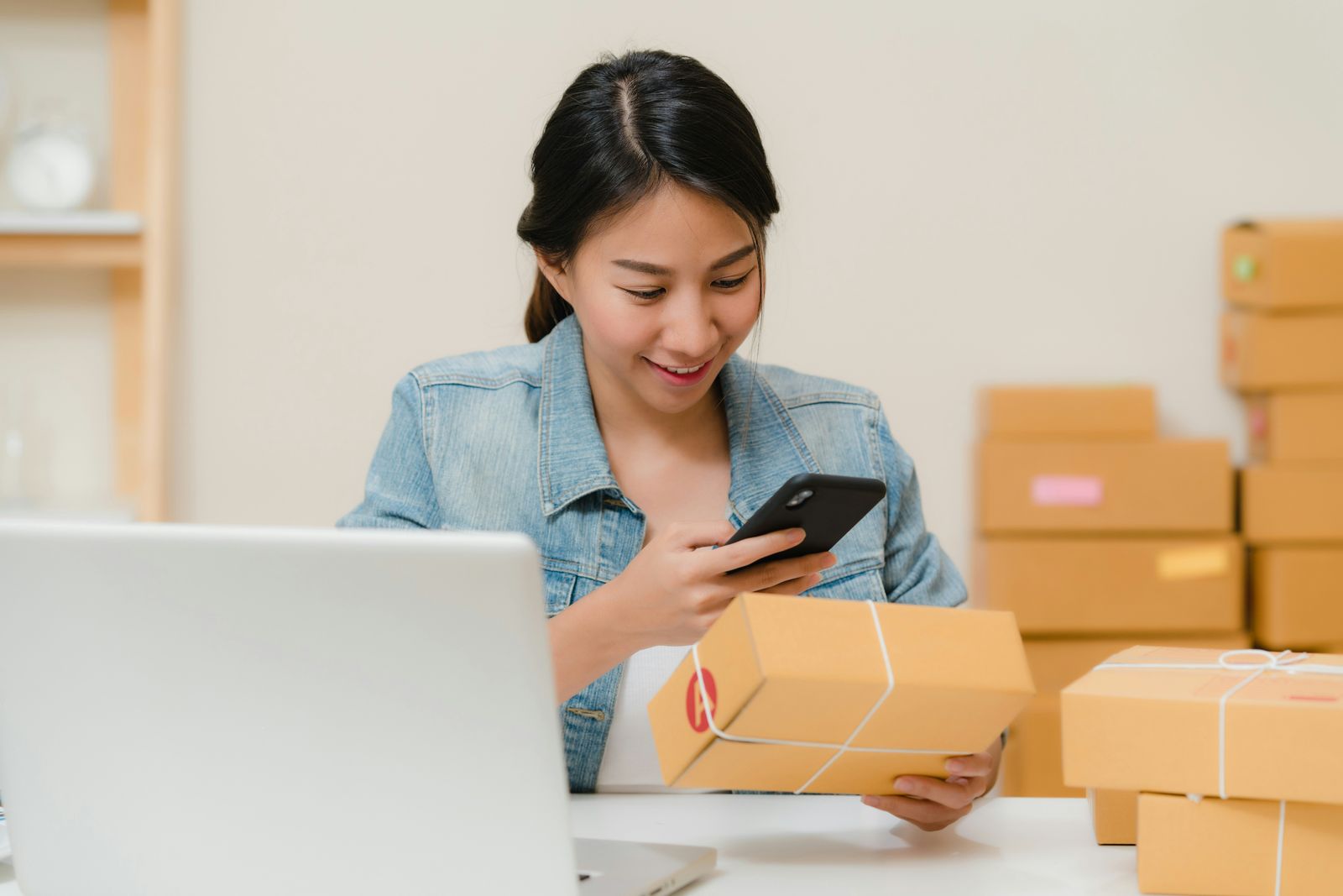 Donna che controlla un pacco con il telefono accanto al portatile e alle scatole di un negozio online